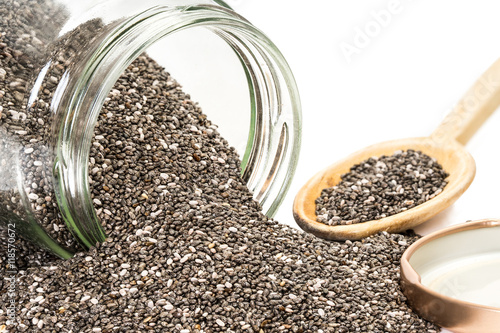 Chia seeds coming out of an overturned glass jar with the lid next to it and a wooden spoon in the background isolated on white