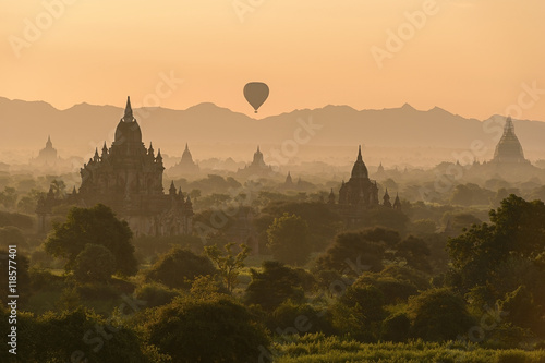A colorful hot air balloon floats over a lush green forest  with  temples peeking through the trees.  In the distance  there are  mountain ranges.