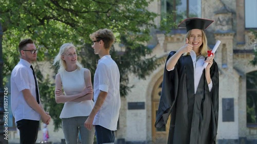 Graduand Woman in Mantle Holding Diploma Smiling Shows Her Thumb Standing in Alley Outside the University Happy Graduands Behind the Woman Sunny Day photo