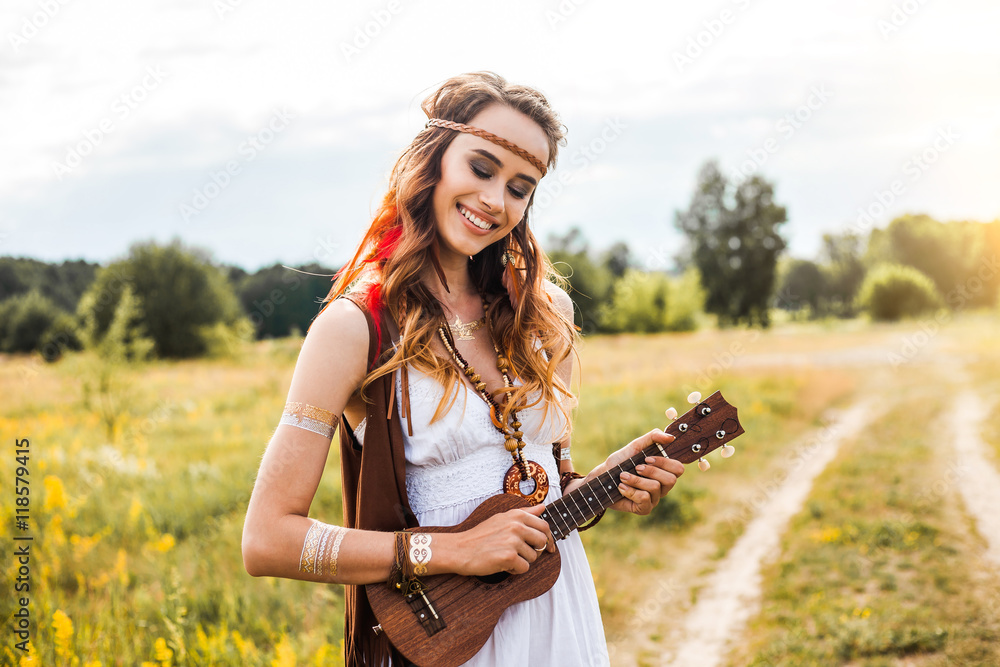 Pretty amazing free red-haired hippie girl playing a ukulele outdoors,  feathers and braids in her hair, white dress, leather and gold accessories,  flash tattoo, indie, Bohemia, boho style, nice smile Stock Photo |