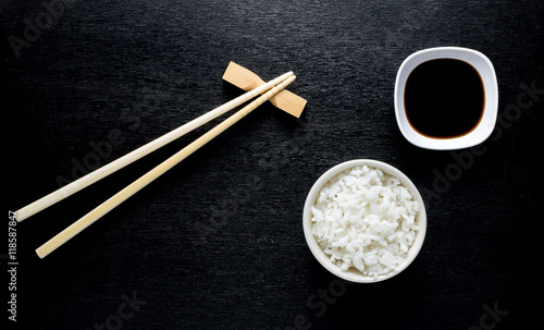 Japanese sushi chopsticks over soy sauce bowl, rice on black bac