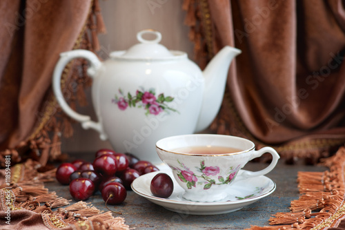 Вишневый чай в изящной чашке на фоне бархатных штор. Рядом с чайным сервизом лежат  ягоды вишни.