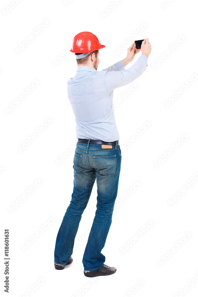 Backview of business man in construction helmet stands and enjoys tablet or using a mobile phone. Standing young guy. Rear view people collection. backside view of person. Isolated over white Backview of business man in construction helmet stands and enjoys tablet or using a mobile phone. Standing young guy. Rear view people collection. backside view of person. Isolated over white