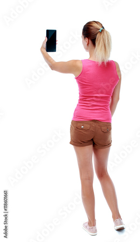 back view of standing young beautiful woman using a mobile phone. girl watching. Rear view people collection. backside view of person. Isolated over white background. Sport blond in brown shorts