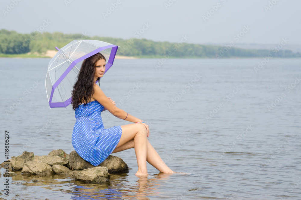 Naklejka premium Lonely sad young woman with an umbrella sits on a rock by the river