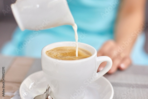 Lady in blue dress pours milk in white cup with coffee