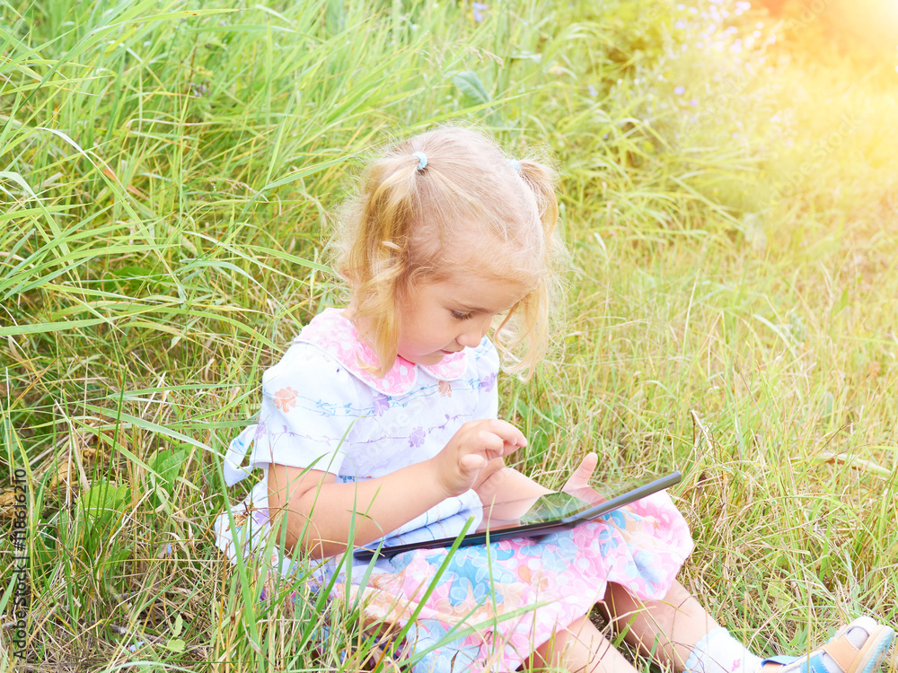 Little girl sitting in park on sunny day and play with tablet pc Little girl sitting in park on sunny day and play with tablet pc