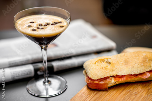 Shakerato drink in cocktail glass with panini and newspaper on the table. Italian lunch with traditional coffee and sandwich with prosciutto photo