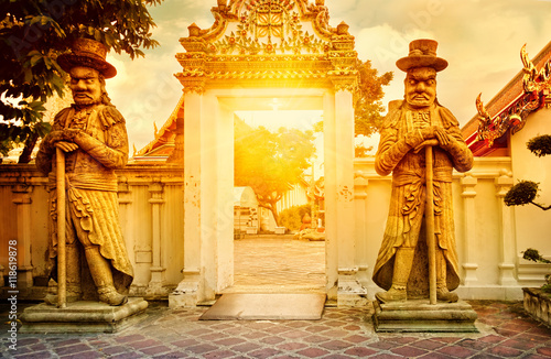 Classical Thai architecture in public temple at dramatic orange sunset sky, Bangkok, Thailand. Guardian Statues stand guard at a gate of Wat Pho. Soft focus.