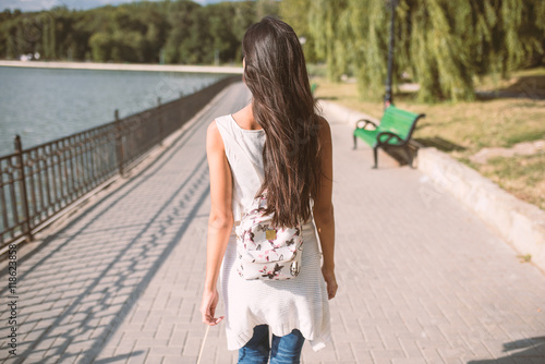Beautiful back view of young brunette woman with long hair and with backpack, walking in the park with lake. Travel. Inspiration © iuricazac