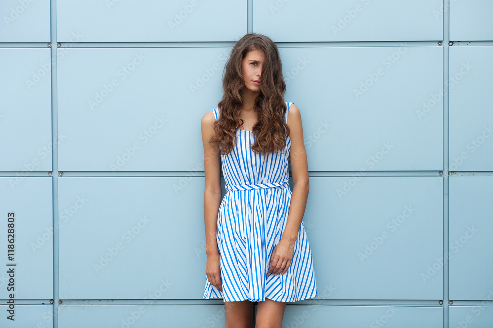 Outdoor portrait of happy young beautiful brunette woman in striped summer dress posing against steelblue metal geometric background Outdoor portrait of happy young beautiful brunette woman in striped summer dress posing against steelblue metal geometric background