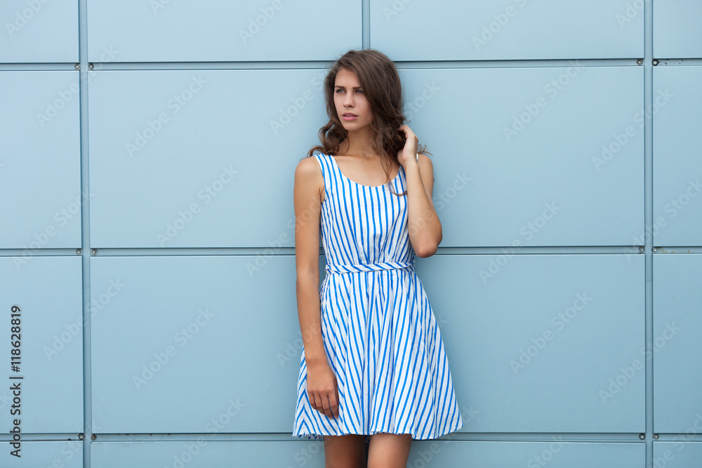 Outdoor portrait of happy young beautiful brunette woman in striped summer dress posing against steelblue metal geometric background Outdoor portrait of happy young beautiful brunette woman in striped summer dress posing against steelblue metal geometric background