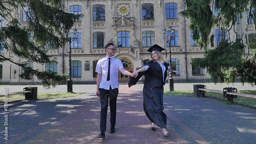 Couple of Graduands Smiling Holding Hands Woman is Twisting Girl in Mantle Guy in Shirt and Tie Walking in Alley Outside the University Happy People photo