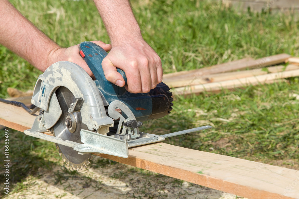 Carpenter hands using electrical powered circular saw to cut wooden plate at the construction site.