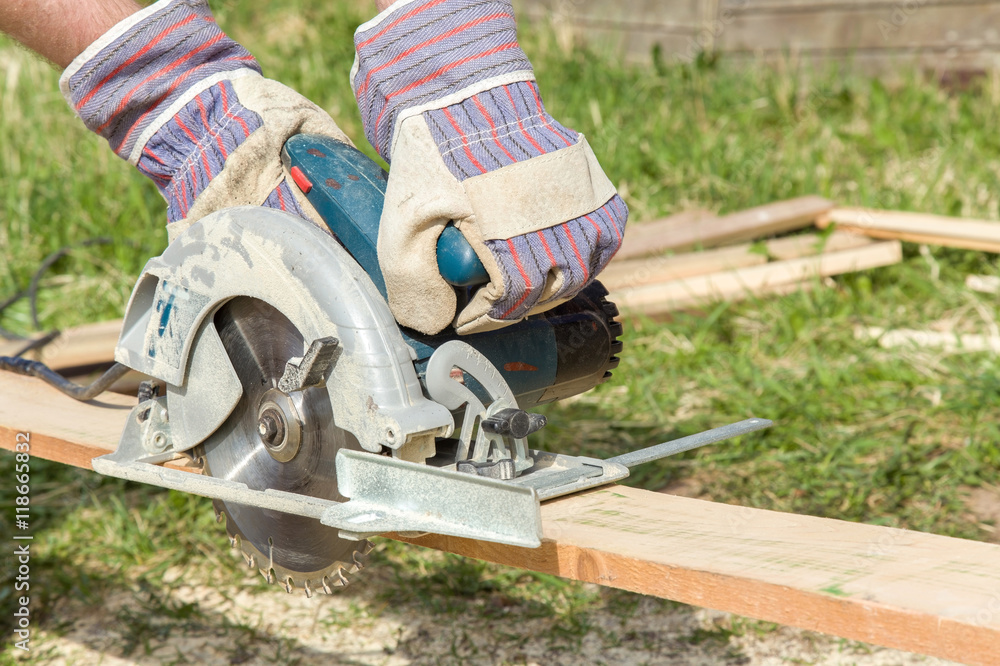 Carpenter hands in protective gloves using electrical powered circular saw to cut wooden plate at the construction site. Carpenter hands in protective gloves using electrical powered circular saw to cut wooden plate at the construction site.