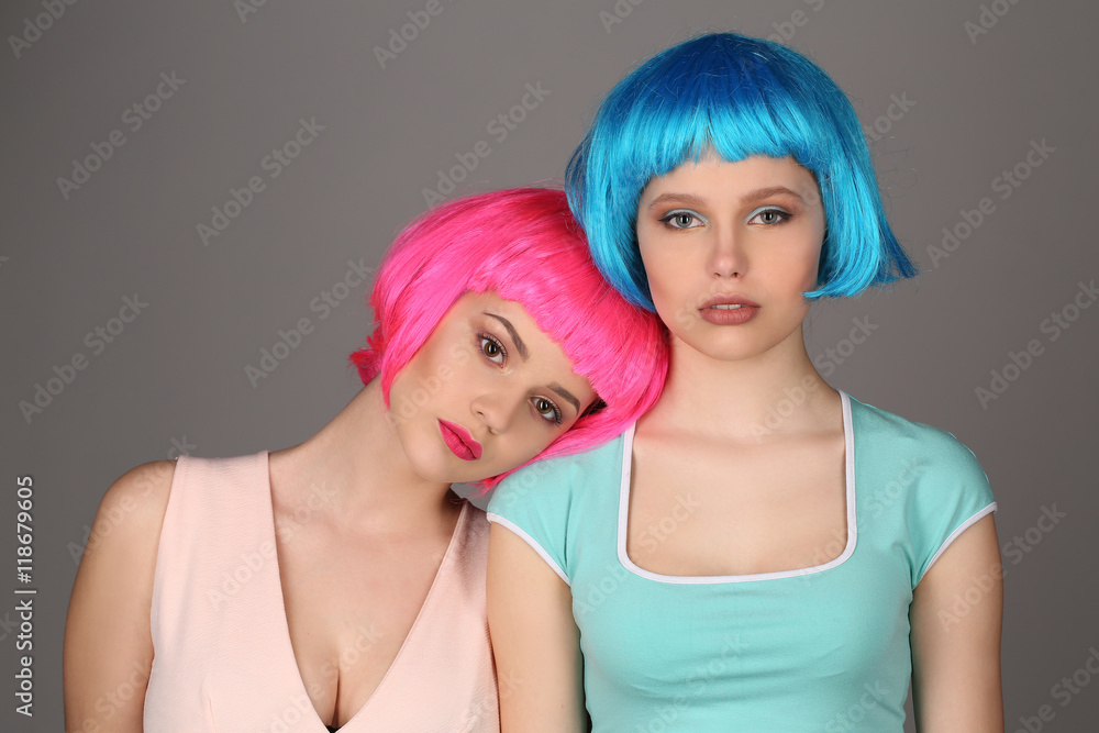 Two girls in colorful wigs standing together. Close up. Gray background