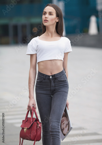 Beautiful brunette young woman wearing black jeans, white shirt and walking on the street . Fashion magazine in hand. Blurred background.