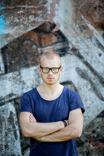Portrait of the young man in glasses against the background of a concrete wall with graffiti. The guy looks at the camera.