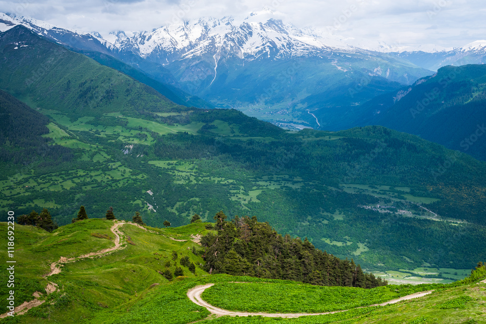 Mountain panorama with a green field slope, wooded mountains and snowbound peaks in the background. Hairpin bends of a dirt road. Svaneti, Georgia. The Caucasus mountains.
