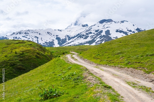 Dirt road in mountains. Road goes through summer pasture highland full of green grass. The foot of Ushba mountain. Snow-capped mountains in the background