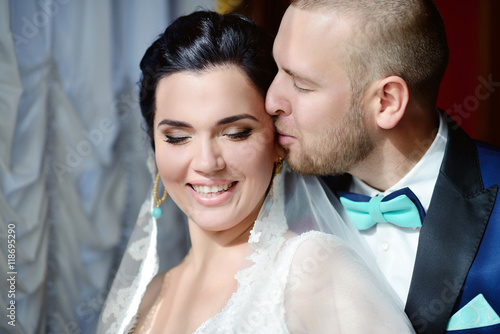 Wedding couple indoors is hugging each other. Beautiful model girl in white dress. Man in suit. Beauty bride with groom. Female and male portrait. Woman with lace veil. Cute lady and handsome guy