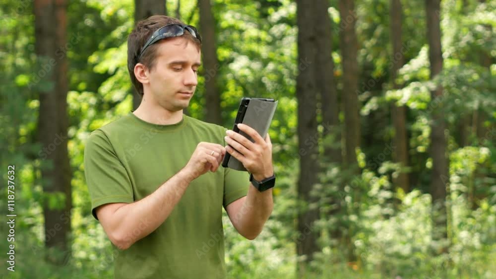 Young man touching tablet in the park among the trees. Green T-shirt and sunglasses Young man touching tablet in the park among the trees. Green T-shirt and sunglasses