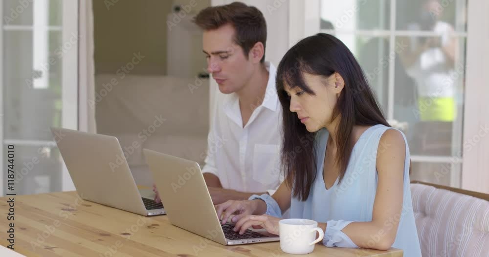 Calm young couple in loose summer clothing sitting at wooden table using similar laptop computers. Includes copy space. Calm young couple in loose summer clothing sitting at wooden table using similar laptop computers. Includes copy space.