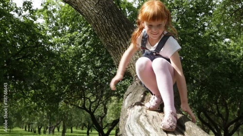 Little ginger girl in a denim dress is climbing down a tree