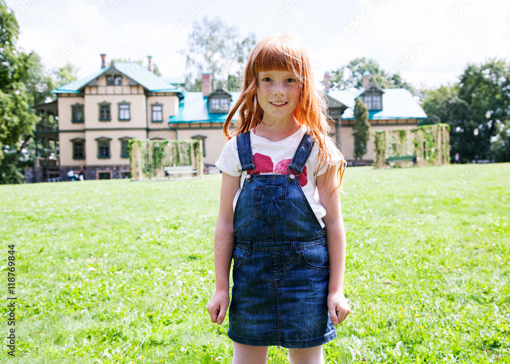 Cute little girl standing against a green lawn and big village house and smiling - obrazy, fototapety, plakaty 