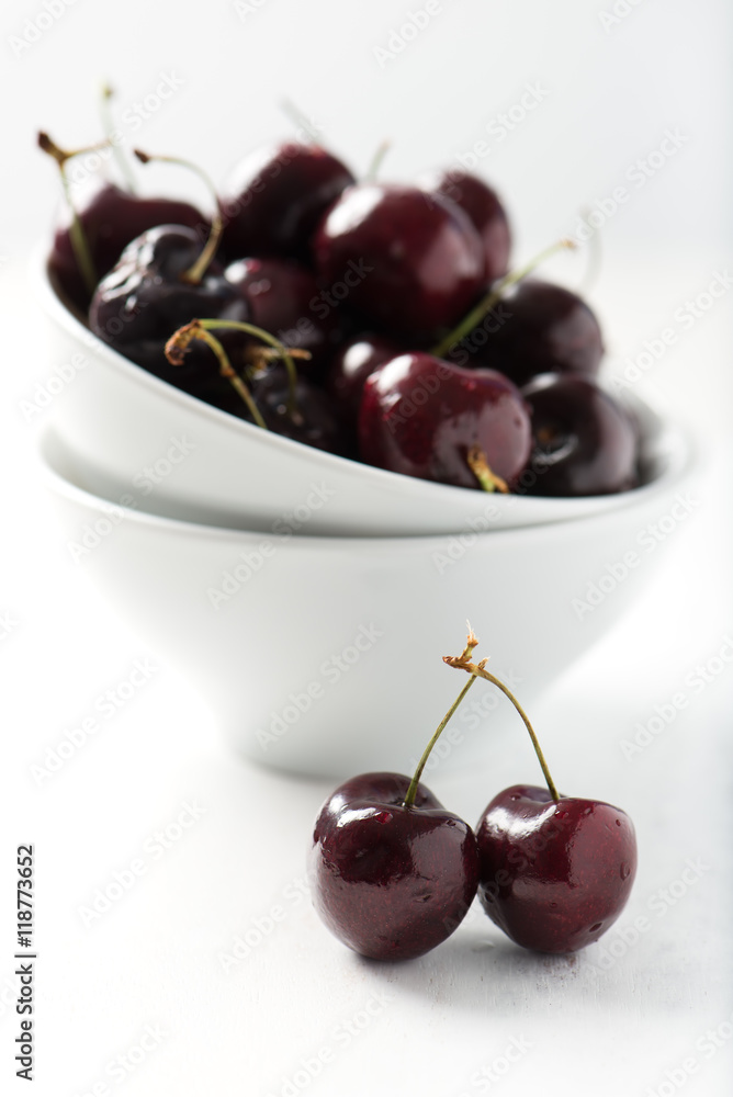 brightly lit, fresh, organic cherries in white bowl, couple of cherries in selective focus at front, isolated on white wooden background, vertical brightly lit, fresh, organic cherries in white bowl, couple of cherries in selective focus at front, isolated on white wooden background, vertical