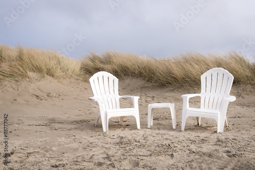 two, white and empty adirondack chairs and coffee table between them over sand dunes on a beach, cloudy, dramatic sky in the background