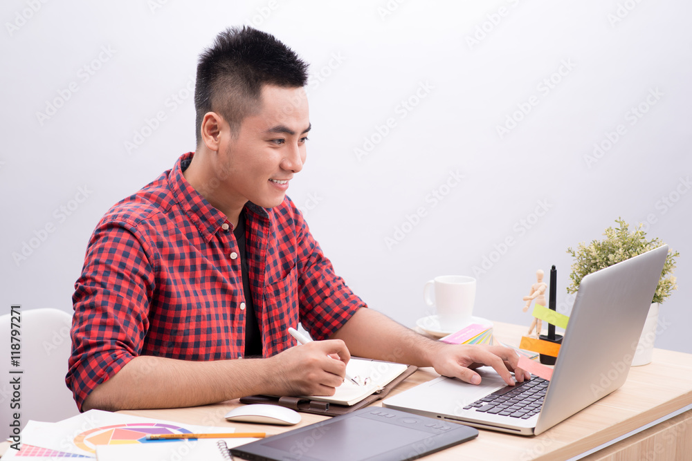 Asian Man Writing At Desk In Busy Creative Office Asian Man Writing At Desk In Busy Creative Office