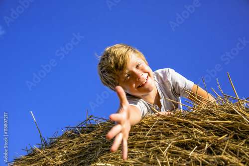 Smiling boy on a haystack on the background of blue sky pulls the arm to the camera