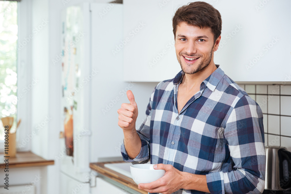 Cheerful young man standing on kitchen and showing thumbs up Cheerful young man standing on kitchen and showing thumbs up