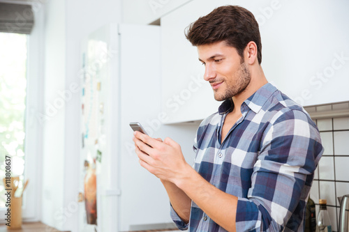 Handsome young man standing and using smartphone on the kitchen