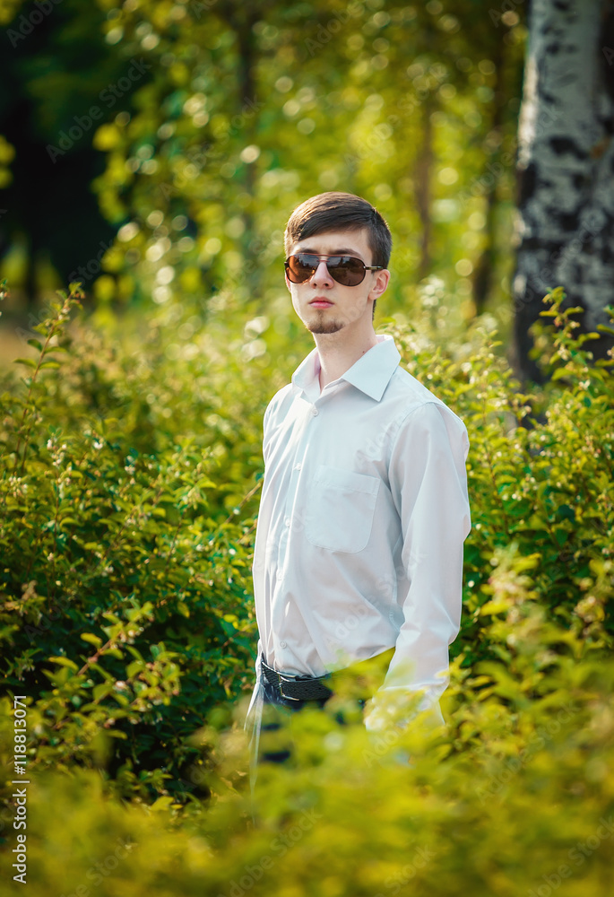 Handsome stylish young man with beard in sunglasses and white shirt with long sleeves standing in the park among green bushes