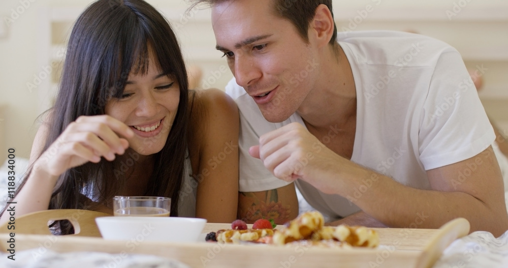 Cute young newlywed couple playing with food while having breakfast in bed with tray of pastries and bowl of fruit