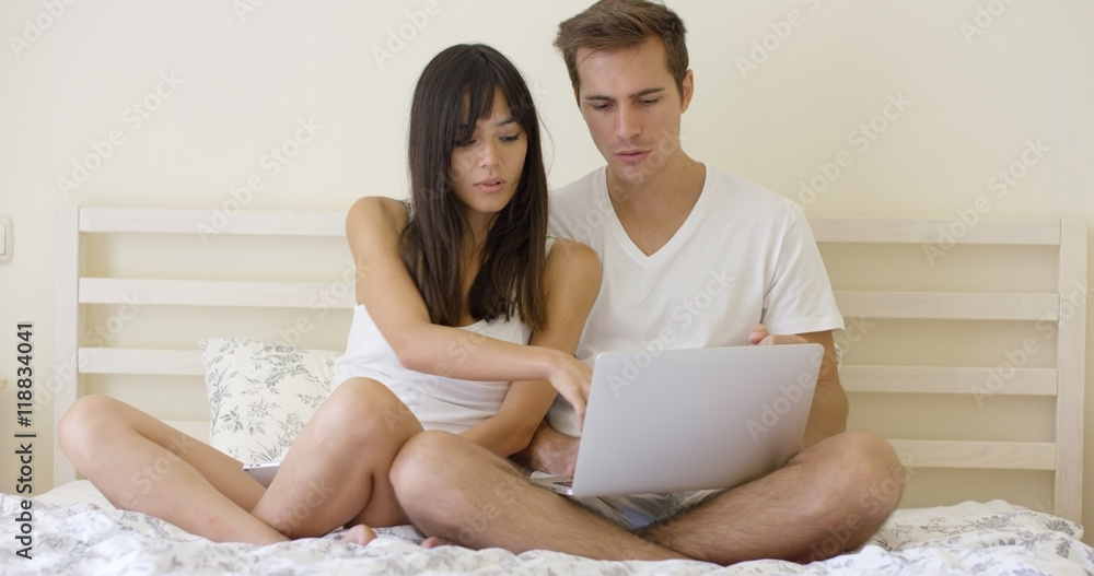 Smiling young long haired woman showing something to handsome man on laptop while in bed Smiling young long haired woman showing something to handsome man on laptop while in bed