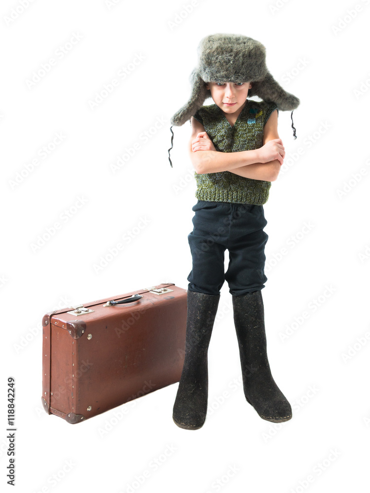 Rustic boy in a fur hat and boots standing near an old suitcase with his hands crossed on a white background Rustic boy in a fur hat and boots standing near an old suitcase with his hands crossed on a white background