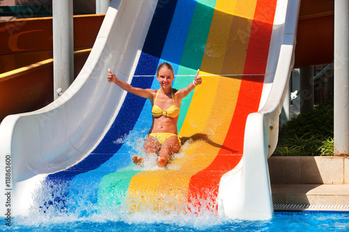 Girl on water slide at water park show thumb up in two hand . Water slides with flowing water in water park.