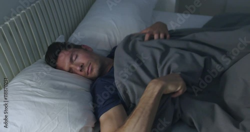 Sleeping man turns towards light while dreaming in a double bed with soft ambient light.  Camera move on jib arm.  Side view, medium shot.