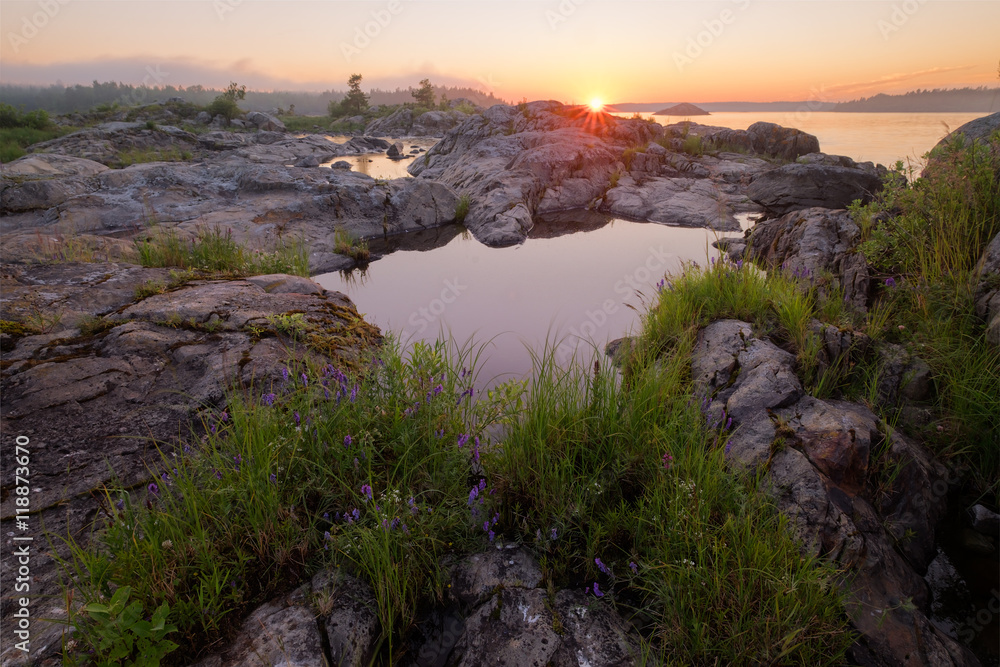 Landscape with small pond covered by granite rocks, surface of the water reflects sun rays of sunset, Russia, Karelia