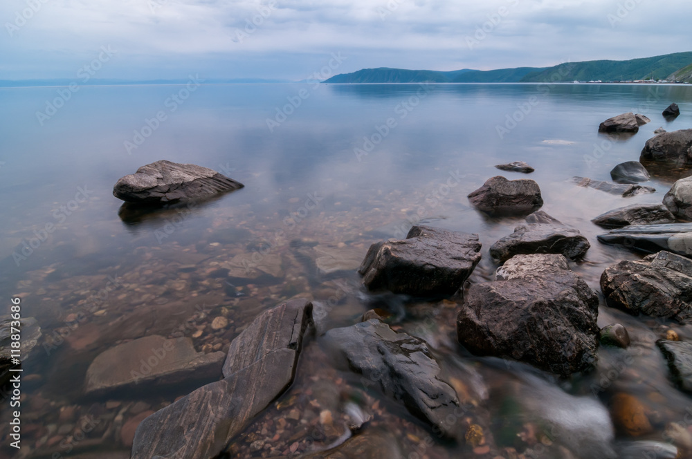 Small creek flow into smooth surface of Baikal lake. Big stones and pebbles seen through clear water, Russia, Siberia