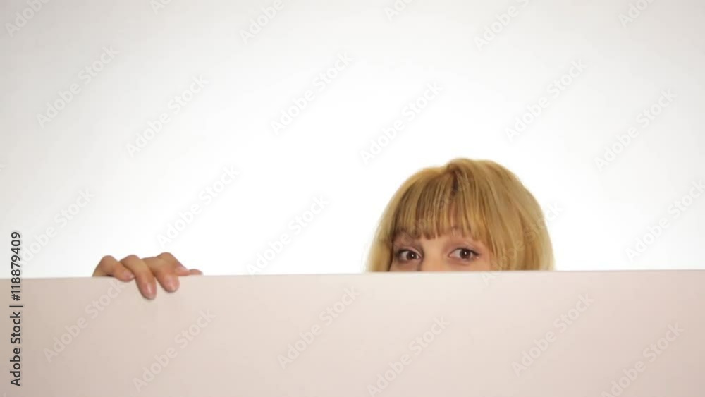 Smiling happy woman standing behind and leaning on a white blank billboard or placard, looking over the edge and hiding behind a placard. over white background Smiling happy woman standing behind and leaning on a white blank billboard or placard, looking over the edge and hiding behind a placard. over white background