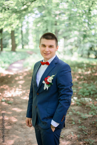 Fiance in blue suit and red bow tie stands on the path in park