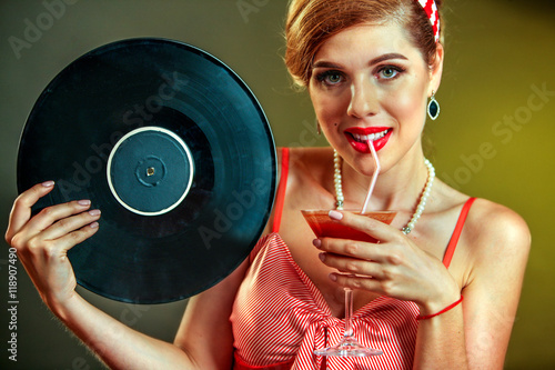 Girl in pin-up style keeps vinyl record and drink martini cocktail . Pin-up retro female style. Girl pin-up style wearing red dress