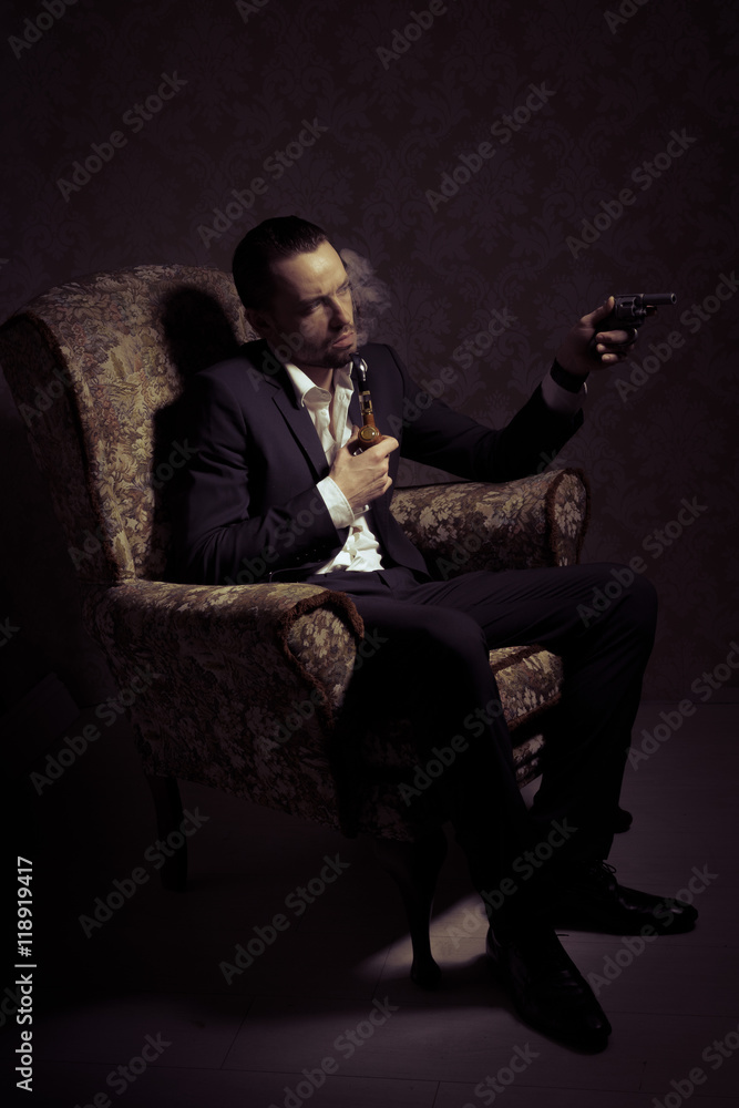 Young handsome and elegant man sitting in chair, holding gun and smoking a pipe isolated over vintage background Young handsome and elegant man sitting in chair, holding gun and smoking a pipe isolated over vintage background