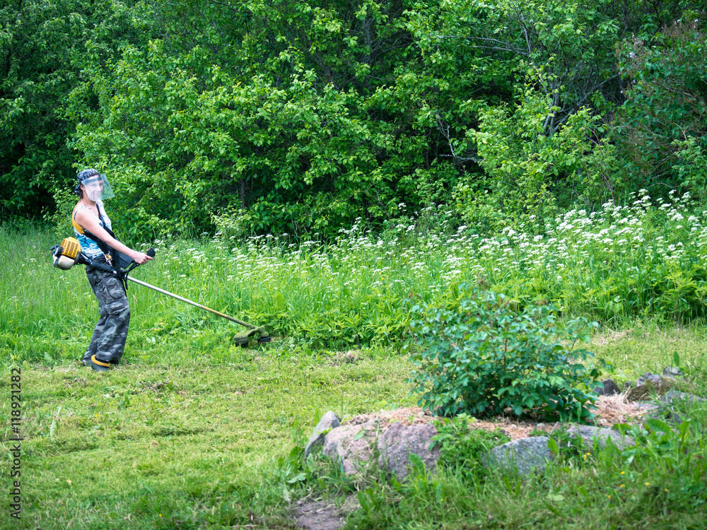 Adult woman in a protective mask holding a streamer and mowing grass against the backdrop of deciduous trees Adult woman in a protective mask holding a streamer and mowing grass against the backdrop of deciduous trees