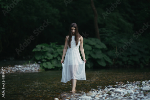 Fine art outdoor portrait of beautiful young woman in a white dress. Girl walking alone by the river.