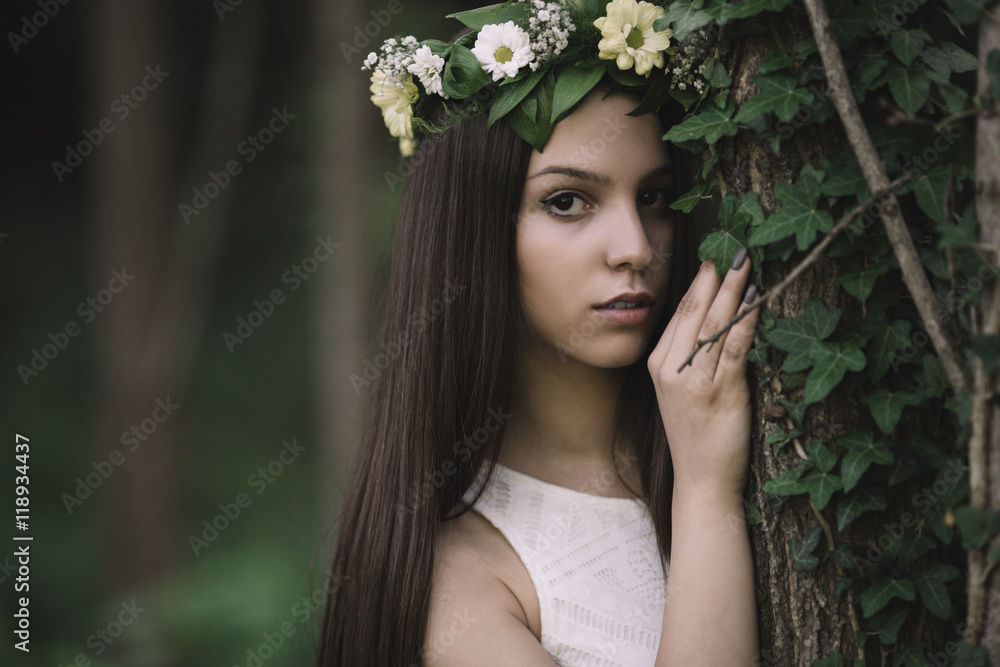 Fine art outdoor portrait of beautiful young woman in a white dress wearing floral wreath.